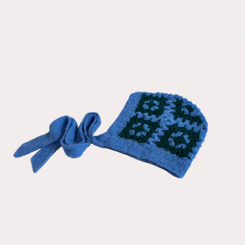 Handgehäkelte Granny Square Bonnet Haube in Hellblau und Dunkelgrün. Die flauschige Häkelmütze ist auf weißem Grund dargestellt, wobei die beiden hellblauen Bindebänder für das Foto auf der linken Seite arrangiert wurden. Sie zeigt das Muster von der Seite.