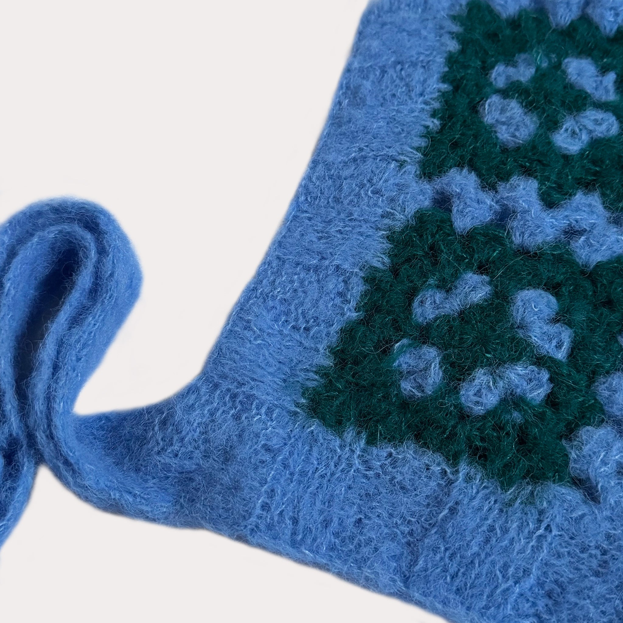Nahaufnahme der Textur einer handgehäkelten Bonnet aus hellblauem Mohair-Mischgarn. Man sieht die weichen Fasern und das detaillierte Granny-Square-Muster in Dunkelgrün. Am Rand ist der Ansatz eines schmalen, hellblauen Bindebandes zu erkennen.