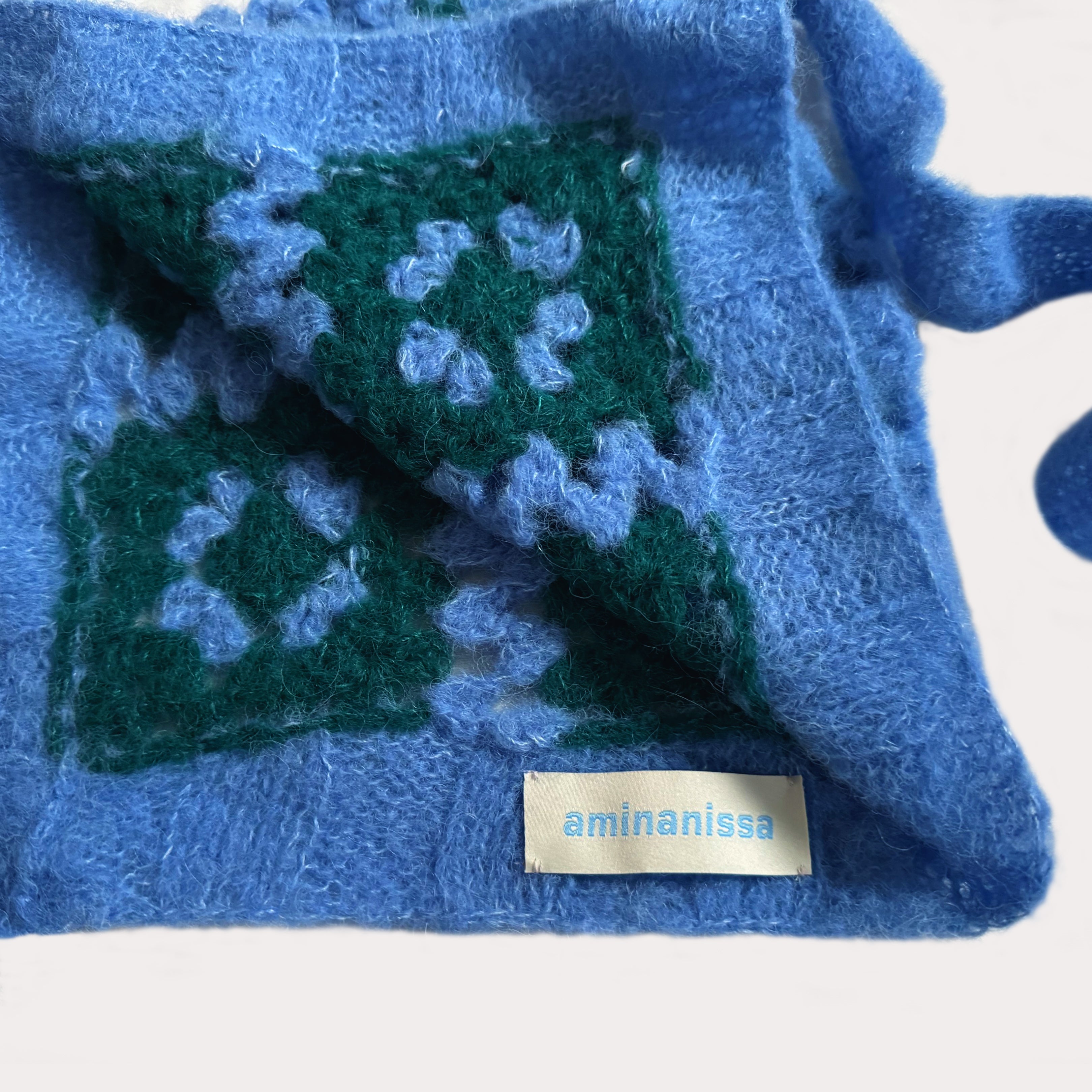 Nahaufnahme einer hellblauen Granny Square Bonnet mit dem weißen Marken-Label „aminanissa“. Man sieht die weiche, flauschige Textur der Häkelarbeit und die hochwertige Verarbeitung der dunkelgrünen Details auf weißem Grund.