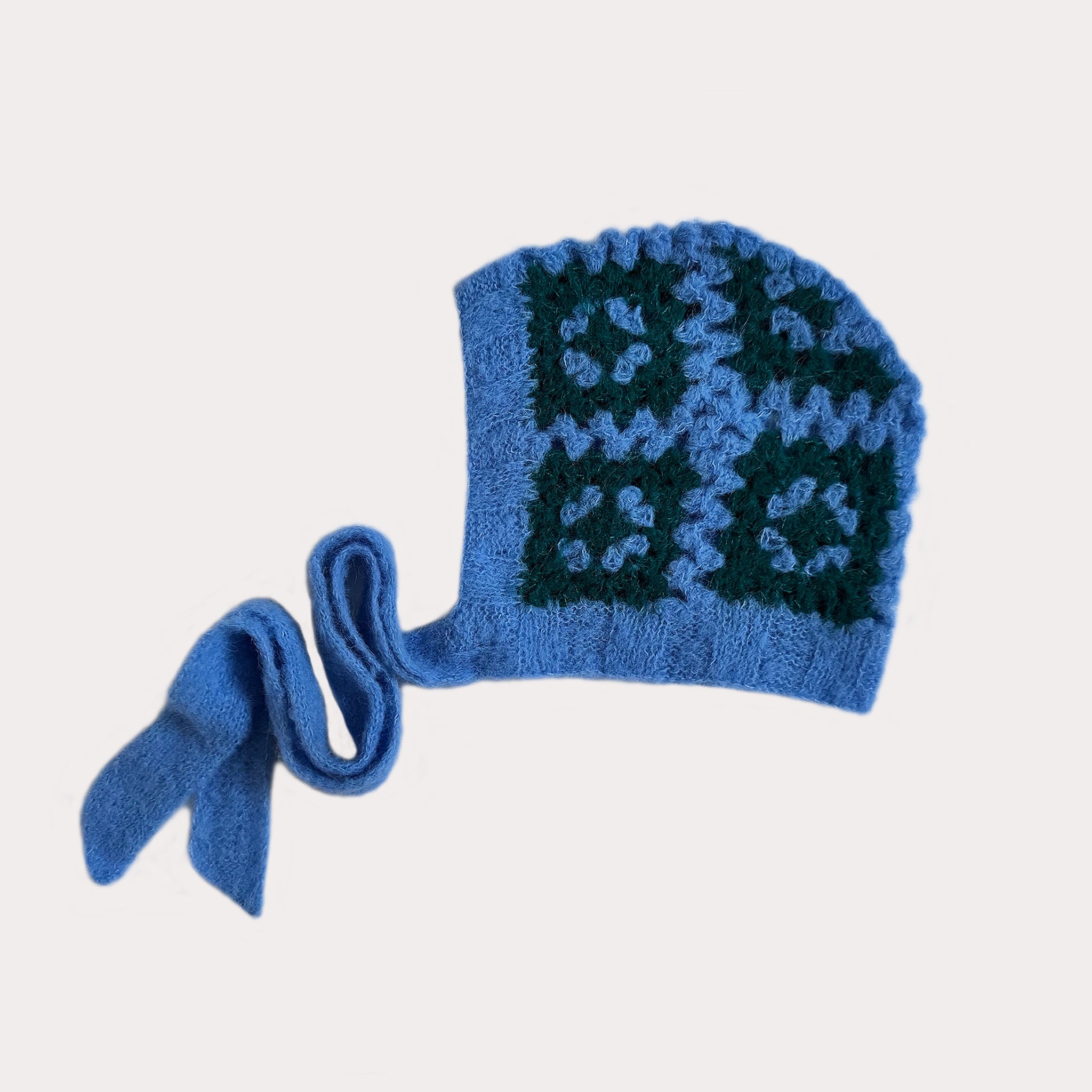 Eine handgehäkelte, flauschige Bonnet-Haube aus leuchtend hellblauem Garn, fotografiert auf einem weißen Hintergrund. Die Mütze zeigt ein Muster aus vier Granny-Squares in Dunkelgrün mit hellblauen Akzenten. Von der Unterseite der Haube führen zwei schmale, hellblaue Bänder ab, die auf diesem Bild dekorativ auf der linken Seite abgelegt sind. Die Haube wird im Profil von rechts gezeigt.